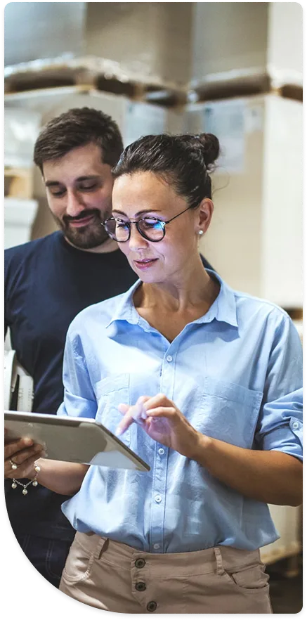 Two colleagues in a warehouse reviewing information on a digital tablet, with shelves and boxes in the background.