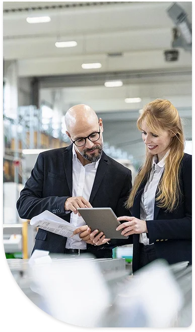 Two professionals, a man and a woman, in formal attire, reviewing information on a tablet in a modern warehouse environment.